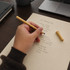 A hand holding a tan-coloured inkless pen, writing on a cream coloured notepad, with a coffee mug and laptop in the background.
