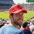 A man wearing a red Classic 5 Panel Trucker Mesh Cap smiles at a sports event, with a crowd in the background.
