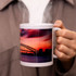 A white sublimation mug featuring a scenic design with a sunset and bridge, held by an individual with steam rising.