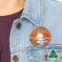 A 65mm button badge with a sunset design, featuring warm colours, attached to a denim jacket. It includes an Australian Made logo.