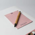 A pink notepad with lined pages and a wooden pen resting on top, featuring a logo.