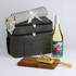 A cooler bag in grey with a blanket, a bottle of white wine, a wooden cutting board, and gold utensils.