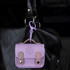 A small, lavender mini tech charm case with a clasp hanging from a black handbag. It has a decorative design and two tan straps.