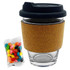 A glass coffee cup with a black lid and cork band, accompanied by a packet of colourful jelly beans.