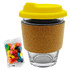 A glass coffee cup with a cork band and a yellow lid, accompanied by a pack of colourful jelly beans.