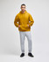 A mustard yellow pullover hooded sweatshirt worn by a model with grey track pants and black shoes. The sweatshirt has a logo.