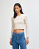 A fitted long-sleeve crop t-shirt in a light beige colour, paired with blue jeans. The model is standing and looking directly at the camera.