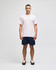 A man stands wearing a light pink short sleeve t-shirt and navy shorts, featuring a logo on the t-shirt.
