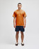 A man stands in a burnt orange short sleeve t-shirt paired with navy shorts, featuring a logo on the shirt.