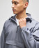 A grey packable anorak jacket with a hood, featuring a front pocket and logo on the cuff.