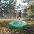 A green frisbee rests on the ground, surrounded by trees and people playing in the background. It features a logo.