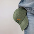 A vintage washed cap in olive green, featuring a small logo, hanging from a denim pocket.