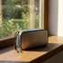 A grey Bluetooth speaker sits on a windowsill, showcasing a sleek design with a logo on its front.