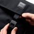 A black messenger bag with a flap and fastening mechanism, featuring a logo. A hand is holding the clasp.