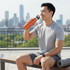 A drink bottle in vibrant orange is being held by a man sitting outdoors on a bench, with a city skyline in the background.