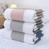 A stack of four blankets in grey, white, pink, and teal, neatly folded on a bed. The image features a logo.