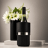 A bottle cooler in black with a silver band, featuring a wine bottle and a vase of flowers in the background.