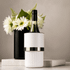 A white wine bottle cooler with a silver band, placed on a marble surface beside a black vase with white flowers.