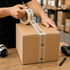 A cardboard box being sealed with transparent packaging tape, featuring a branding logo along the tape.
