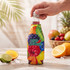 A drink bottle in a vibrant, multi-coloured stubby cooler featuring a fruity design, with a hand holding it.