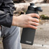 A drink bottle in matte black with a screw-top lid, held by a person sitting outdoors. It features a logo.