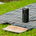 A black rugged fabric waterproof Bluetooth speaker placed on a checkered picnic blanket, set on grass.