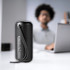 A black rugged fabric waterproof Bluetooth speaker with a handle, positioned on a desk with a blurred person and laptop in the background.