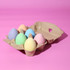 A carton containing six pastel-coloured chalk pieces shaped like eggs, set against a pink background.