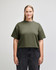 A woman wearing an olive green mid-length boxy top with short sleeves, paired with light blue jeans. The top features a logo.
