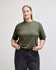 A women's mid-length boxy top in dark green, featuring a relaxed fit and short sleeves. The model is standing confidently.