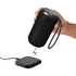 A black outdoor Bluetooth speaker held above a charging pad, featuring a textured finish and a carrying strap.