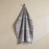 A flat terry towel in shades of grey, hanging neatly with fringed edges. It features a logo on the fabric.