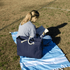 A navy waterproof beach bag with rope handles rests beside a person reading on a blue and white blanket.