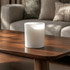 A white scented candle with soy wax sits on a wooden table, surrounded by a softly lit living space.