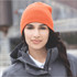 An orange knit beanie worn by a person with long dark hair, paired with a grey jacket. The beanie has a logo.