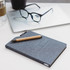 A grey A5 notebook with a textured cover, a wooden pencil, glasses, and a laptop in the background.