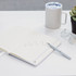 A white A5 notebook with lined pages, a silver pen, a coffee mug, glasses, and a small potted plant on a desk.