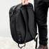 A black backpack with padded straps and a sleek design, held by a person. The bag features a branding logo.