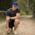 A man wearing a blue Falcon Flex Cap kneels on a sandy path, dressed in a grey shirt and shorts, with a watch on his wrist.