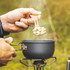 A hand holds a light-colored wooden utensil over a steaming gray pot, set against a natural background.