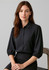 A black 3/4 sleeve blouse with a button-down front and rounded sleeves, worn by a model seated on a chair.