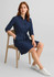 A navy blue dress with two pockets, roll-up sleeves, a belt, and a model seated on a wooden chair.