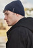 A side view of a man wearing a navy ribbed beanie, paired with a black jacket, outdoors. The beanie features a logo.