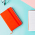 An orange A6 PU notebook with a closure band, beside a white lined notebook on a blue and pink background.