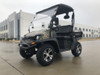 Trailmaster Taurus 200U Utility SXS UTV, rugged off-road vehicle with camouflage pattern, four large tires, and a driver cabin with a clear windshield, parked outdoors on a concrete surface.