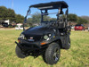 Trailmaster Taurus 200U Utility SXS UTV, black off-road vehicle with four large tires, a spacious interior, and a protective roof, parked on grass with trees and cars in the background.