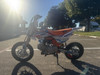 TrailMaster SK110S 110cc Semi-Automatic Kids Dirt Bike, orange and white off-road motorcycle with knobby tires and lightweight frame in an outdoor parking lot under the sun.