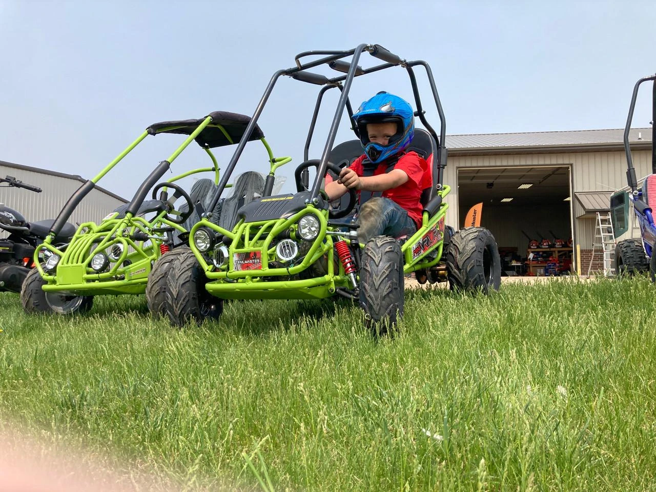 Assembled TrailMaster Mini XRX/R+ Go Kart with Reverse for ages 4 and up, featuring a bright green metal frame, large off-road tires, and a young child wearing a blue helmet sitting in the driver's seat.