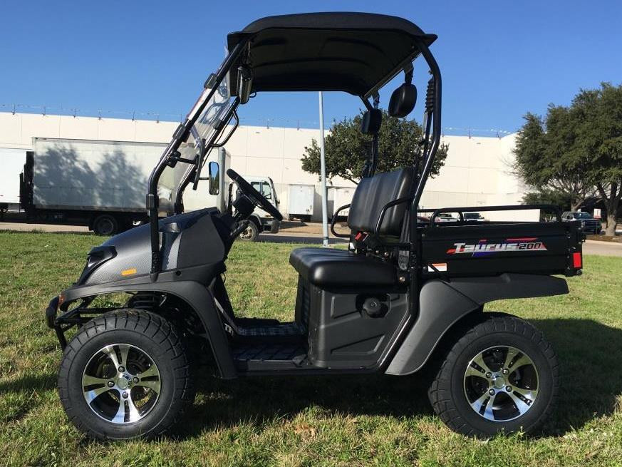 Trailmaster Taurus 200U Utility SXS UTV, black compact utility vehicle with sturdy tires, roof, and cargo bed, parked on grass with clear blue sky.