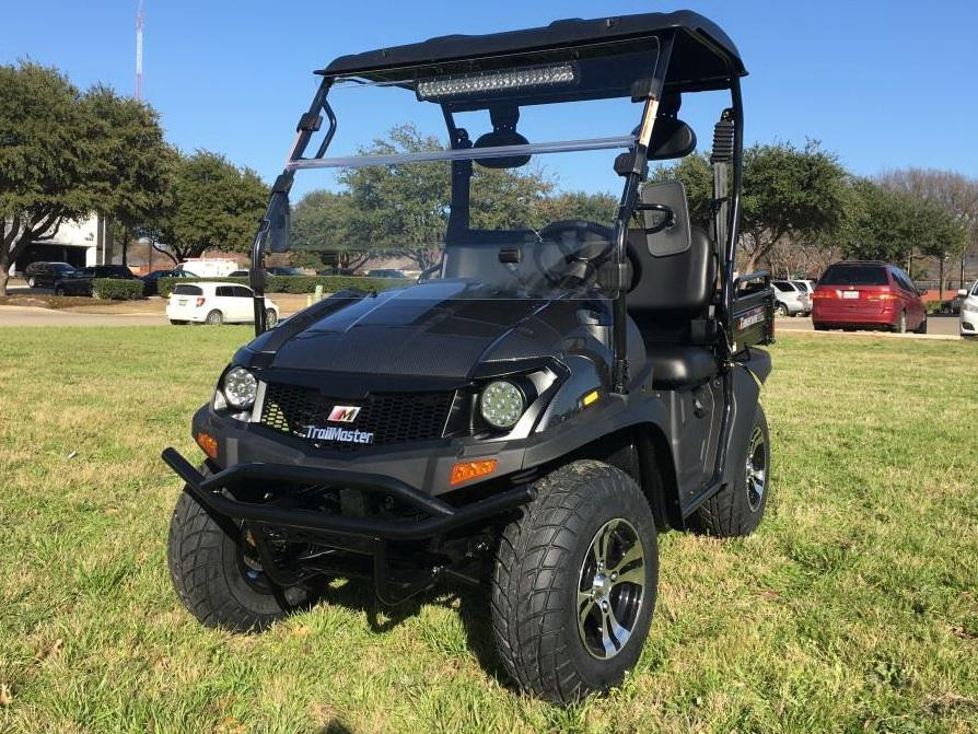 Trailmaster Taurus 200U Utility SXS UTV, black off-road vehicle with four large tires, a spacious interior, and a protective roof, parked on grass with trees and cars in the background.